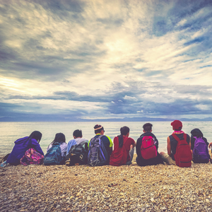 A group of kids wearing backpacks sit on the beach looking out at the sky and water