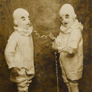 Two creepy old timey children in clown outfits, one holds a string attached to the other