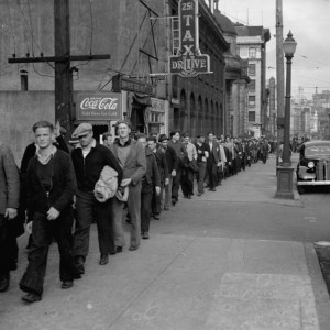 A long line of people stand on a city street in a line for food or work in the 1930s during the Great Depression
