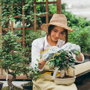 A woman in a sun hat and gloves holds a plant while she sits in a garden