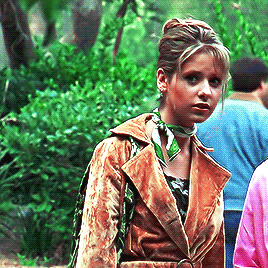 Buffy, rocking an updo and wearing a brown crushed velvet short trench coat and a green scarf tied around her neck