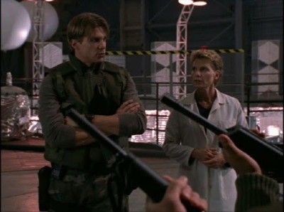 Riley, dressed as a soldier, stands and addresses armed soldiers with Professor Walsh, who wears a lab coat