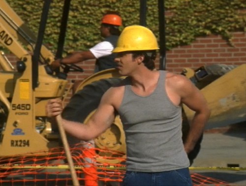 Xander wears a tight tank top, jeans, and a hard hat while working on the construction site