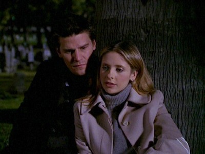 Buffy leans on Angel as they sit against a tree in the nighttime cemetery
