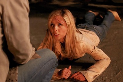 Buffy lays on the ground, wearing a tan jacket, jeans, and heeled boots