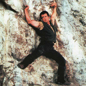 A man dressed all in black (Sylvester Stallone) clings to a cliff face