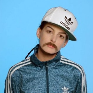 Anne Hathaway dressed in an adidas tracksuit and baseball cap wearing a fake mustache on a blue background.