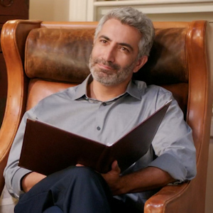 A man with an attentive expression sits on a brown leather chair holding an open note pad