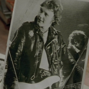 A photo of a young Giles in a leather jacket playing a guitar.