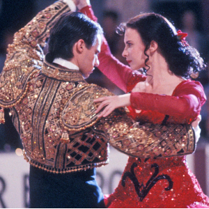 Scene from Strictly Ballroom, with a man in a gold matador jacket dancing with a woman in a red sparkly dress at a ballroom competition