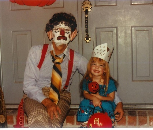 Sarah at age 5, wearing a paper crown and a Mexican dress, sitting next to her dad, who has clown make-up on and is wearing suspenders and a rainbow tie plus a brown wig