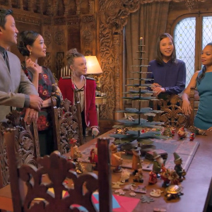 The cast stands around a table with a metal tree with ornaments sitting on it