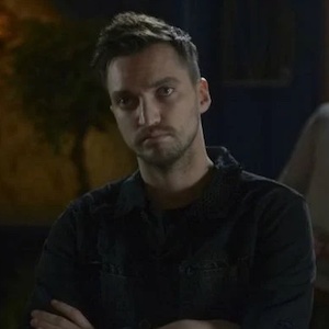 A man in a dark shirt (John Murphy) with his arms crossed glares at someone offscreen 