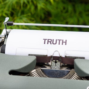 A typewriter with a paper in it with the word truth typed on it sits in front of a blurry background of greenery.