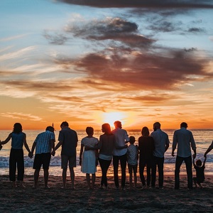 A large group of people holding hands and standing on a beach at sunset, with their backs to the camera