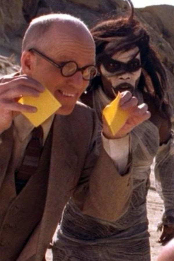 A professorial white man with glasses, holding two slices of cheese and standing next to the original Slayer, whose face is covered in white paint