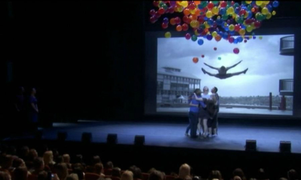A group hugging on stage as balloons drop down, with a photo of Sammy dancing in the background