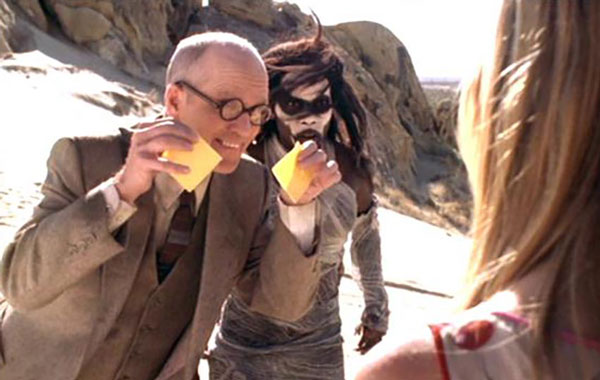 A professorial white guy in glasses, holding up two slices of cheese, next to the original Slayer, her face covered in white paint