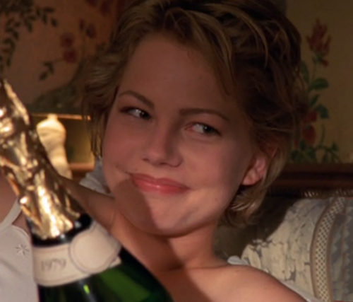 Jen smiles, sitting in bed with a bottle of champagne