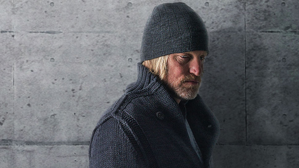 Haymitch, wearing a knit cap and a coat and leaning against a stone wall
