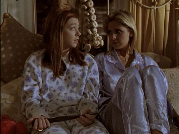 Willow and Buffy sitting on a bed in their pjs with a big string of garlic behind them