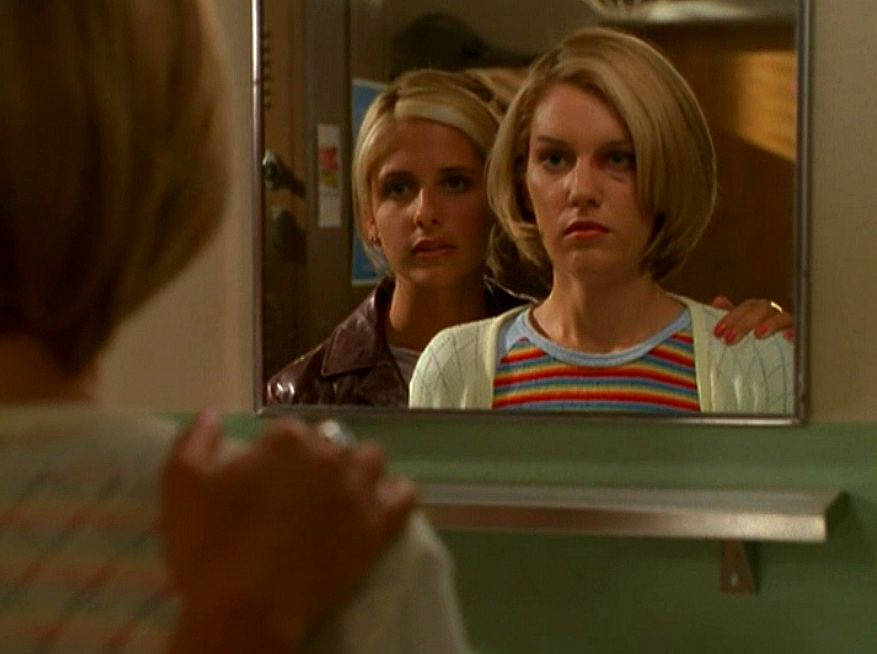 Buffy, standing behind a blonde girl with her hand on her shoulder as they stare into a bathroom mirror