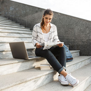 Girl reading a newpaper on steps with backpack and laptop