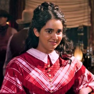 Geraldine Viswanathan in Miracle Workers: Oregon Trail. Viswanathan, in an old timey dress, stands in a saloon