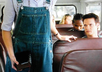 Jamie walking down the aisle of the bus, wearing overalls and carrying a Bible, as Landon looks on in faint disgust