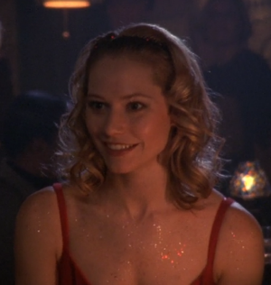 Andie sits, smiling, at the jazz club in a pretty red strappy dress and TONS of body glitter