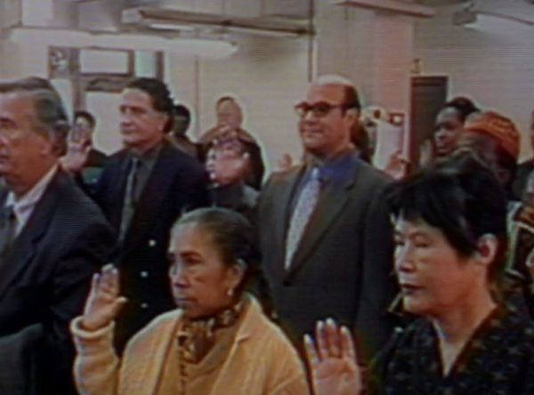 Javier, standing in a room with other people, holding up his right hand as he becomes a U.S. citzen