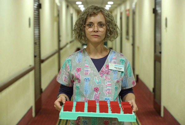 Christina Ricci as Misty, with a bob of blonde curly hair and glasses, wearing scrubs and carrying a tray of pills