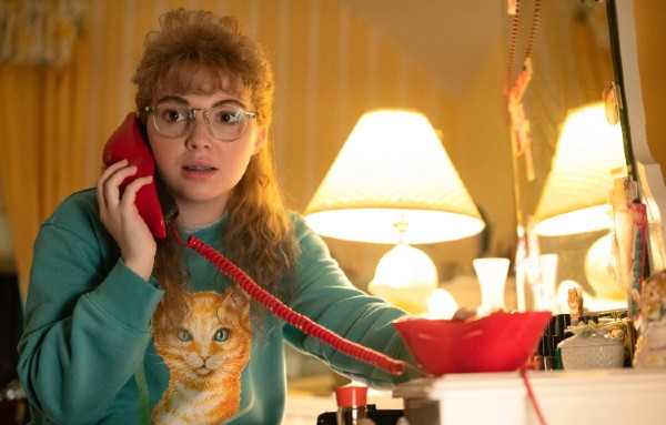 Screenshot from Yellowjackets, with teenage Misty, with big bangs, wearing a cat sweatshirt and talking on a red lip phone