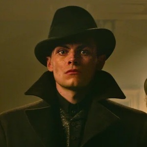 Kaz Brekker from Six of Crows wearing a dark fedora and jacket with a popped collar with a sneer on his face