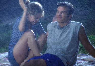 Jen and Jack sit on a blanket on the grass, laughing as they're doused by a sprinkler