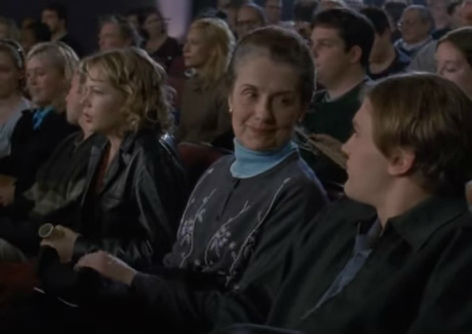 Jen, Grams and Henry sit in a crowd in a darkened auditorium, with Jen staring ahead and Grams smiling at Henry