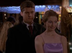 Pacey and Andie watch the dance floor at the prom, Pacey looking serious in a suit and Andie looking amused in a lavender satin gown.