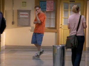 Henry stands in the hallway in a very early aughts outfit of baggie jean shorts, black Chucks and a large orange t-shirt.