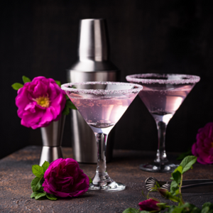 Two martini glasses with pink liquid surrounded by a shaker and flowers on a black background.