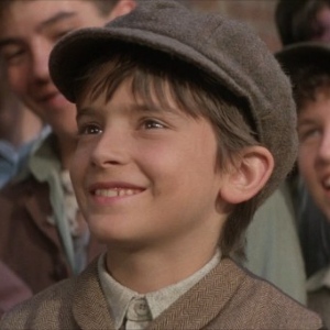 Luke Edwards as Les, a little white kid with brown hair, grinning and wearing a newsies cap