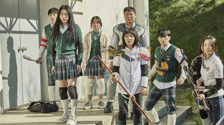 A group of Korean teenagers in uniforms and ragtag protective gear clutching sticks and arrows