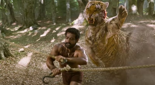A shirtless, wounded Indian man struggles to connect a hook to a rope as a tiger leaps up behind him