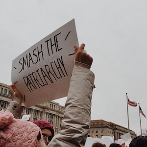 A young woman in a pink hat holds up a protest sign that reads "Smash the Patriarchy"