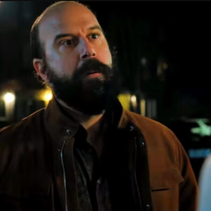 Brett Gelman, a bald man with a dark beard wearing a brown jacket, looks off camera in a disappointed manner