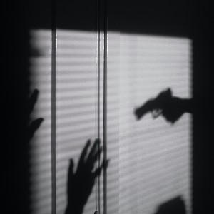 The silhouette of a gun being pointed at someone with their hands up, as seen through window shades