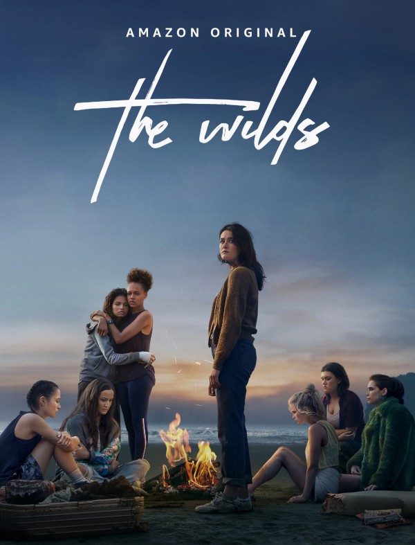 Eight teenage girls sit and/or stand around a bonfire on a beach