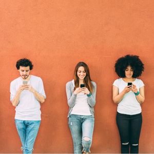 3 people standing against an orange wall looking at their phones.