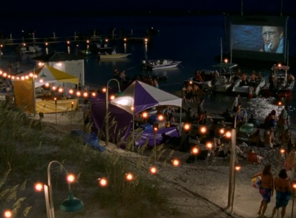 A screening of JAWS plays outside on the water, with kids in boats and on the beach watching it at night.