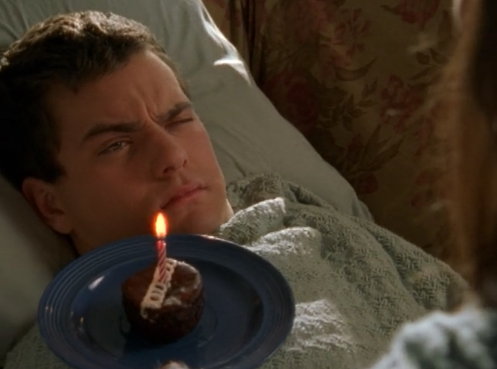 Pacey is bleary-eyed in bed, while a Hostess cupcake with a birthday candle is being held near him by someone off-camera