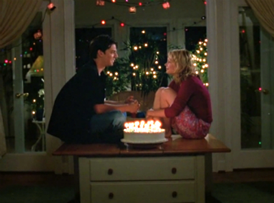 Drue and Jen sit cross-legged in front of a window with a birthday cake lit with candles between them, like the iconic scene from John Hughes' Sixteen Candles